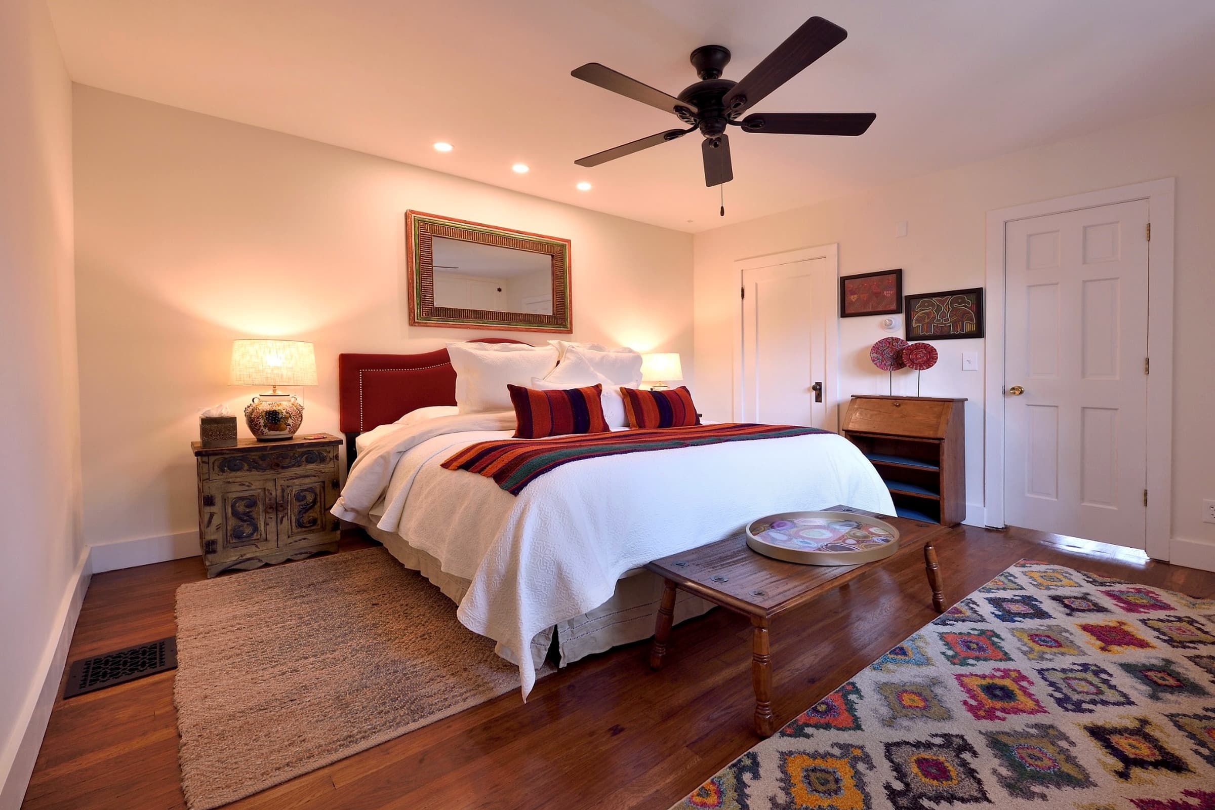 A cozy, well-decorated bedroom featuring a large bed, warm lighting, and a fan.