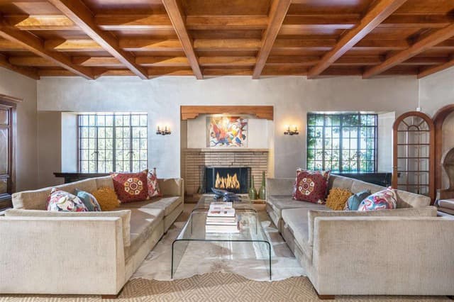 A cozy living room featuring two beige sofas, a glass coffee table, a fireplace, and large windows.