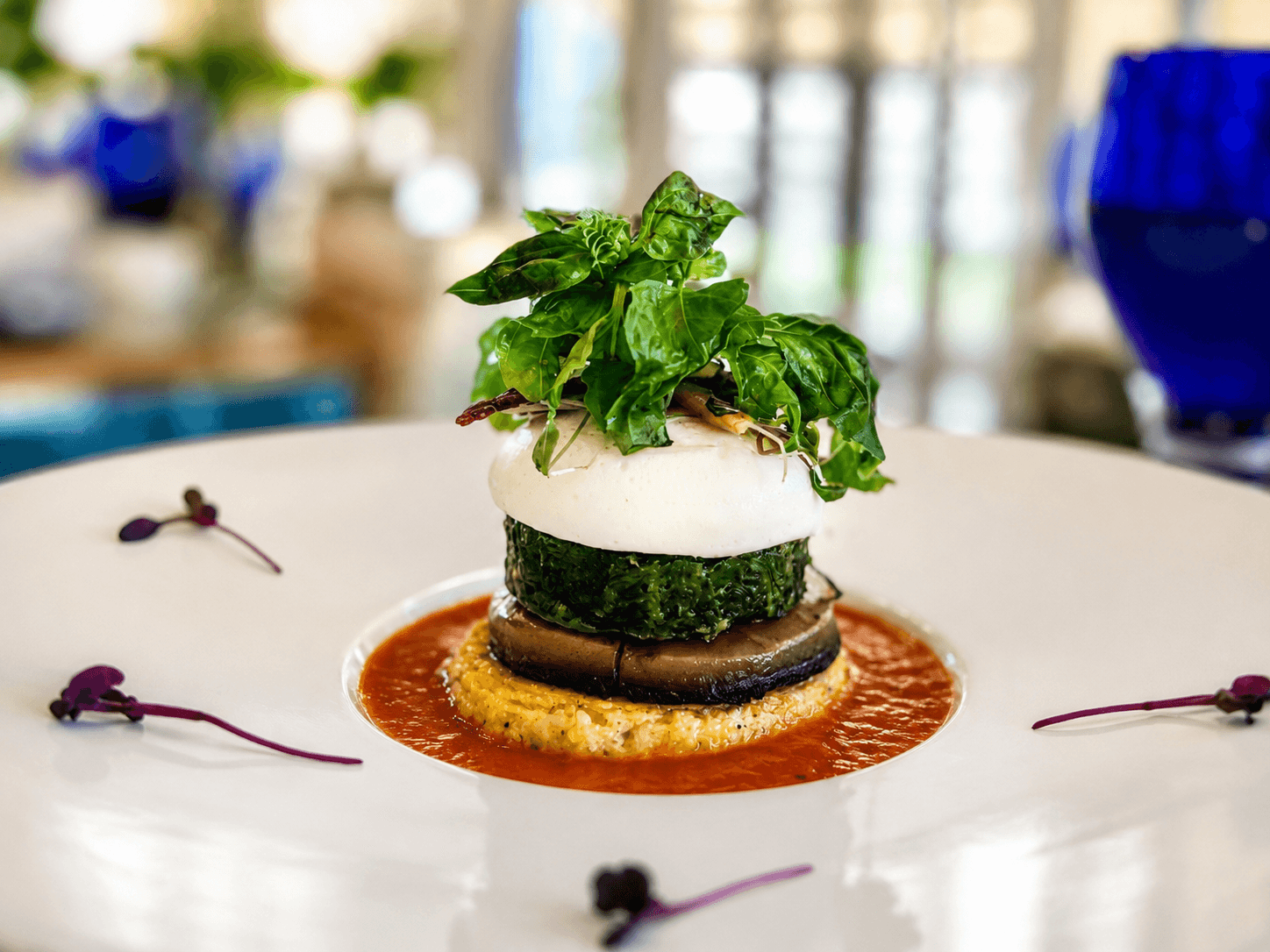 A gourmet dish featuring layered vegetables, topped with greens and served on a vibrant sauce.