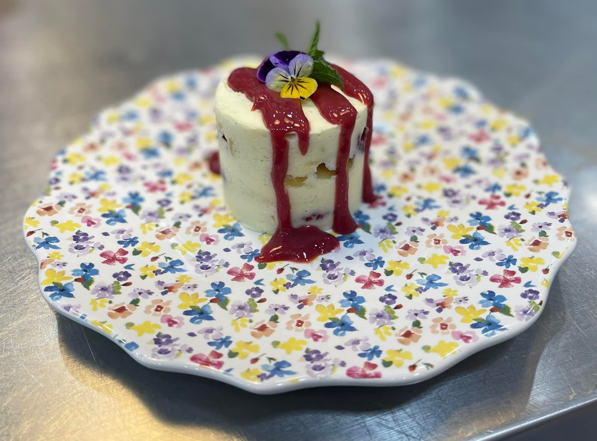 A layered dessert topped with berry sauce and garnished with an edible flower, served on a floral-patterned plate.