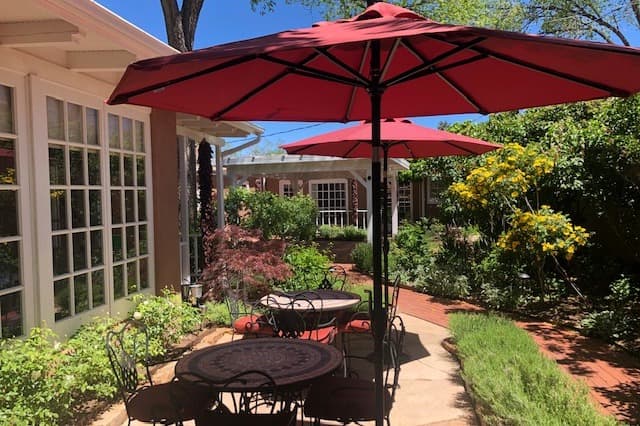 A peaceful outdoor garden area featuring two round tables under red umbrellas surrounded by greenery.