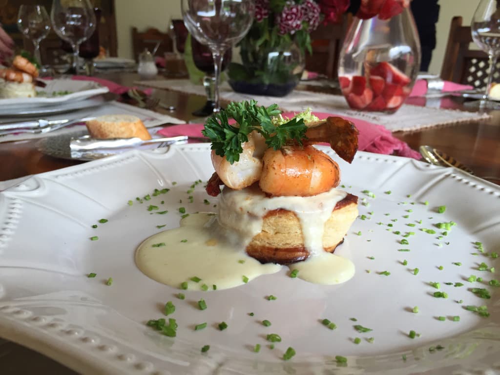 A plated dish featuring a biscuit topped with shrimp, sauce, and herbs, with a dining table set in the background.