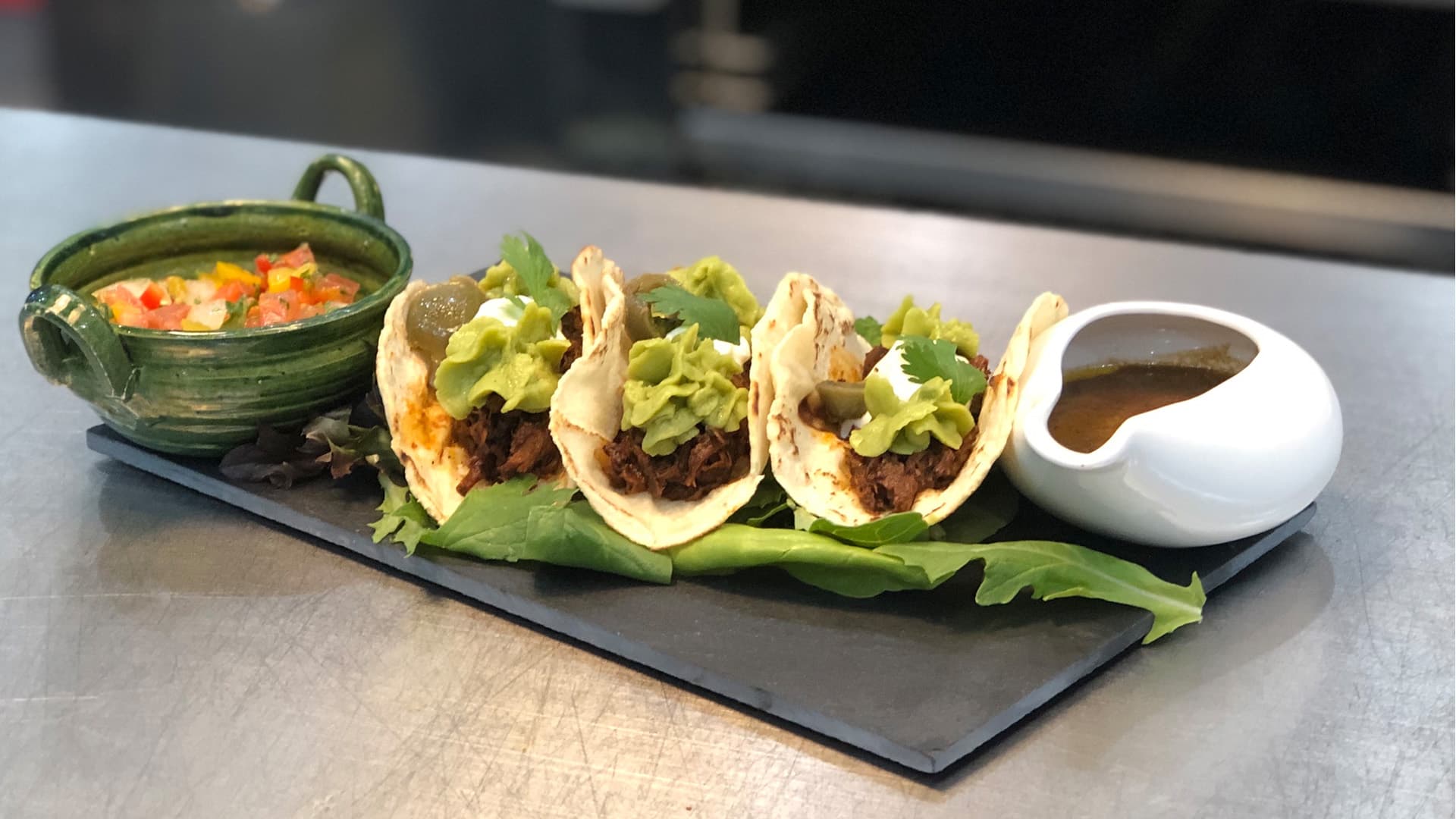 A plate of three tacos filled with shredded meat, topped with lettuce, alongside a small bowl of salsa and a dish of sauce.