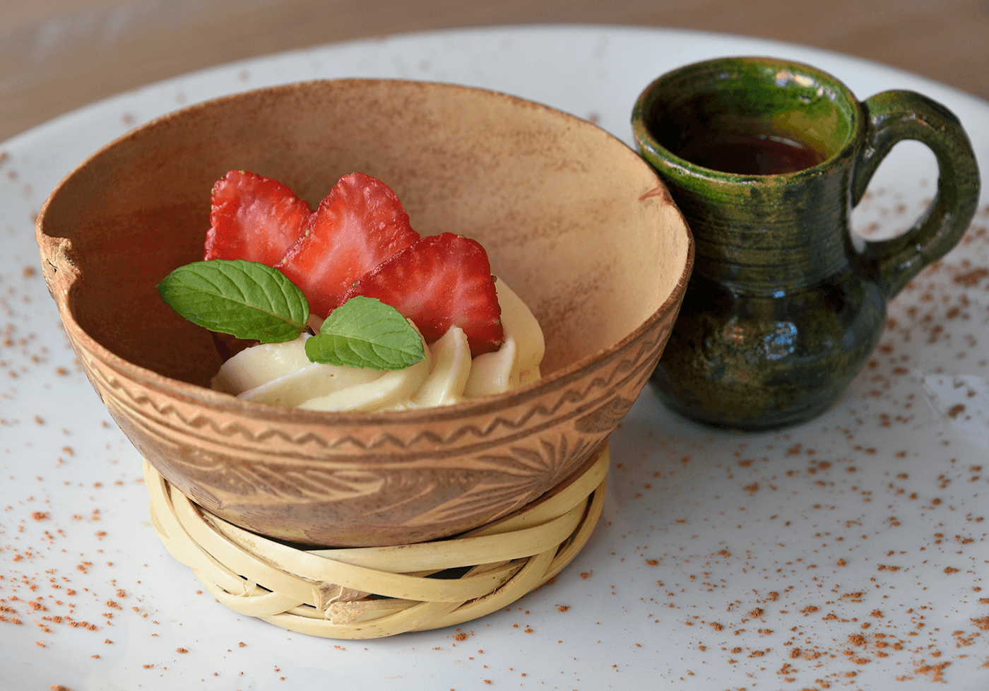 A clay bowl filled with creamy dessert, topped with strawberries and mint, alongside a small green ceramic jug.