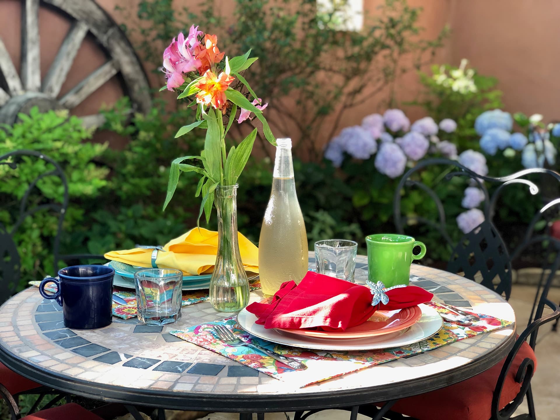 A beautifully arranged outdoor dining table with colorful dishes, glasses, and a vase of flowers surrounded by lush greenery.