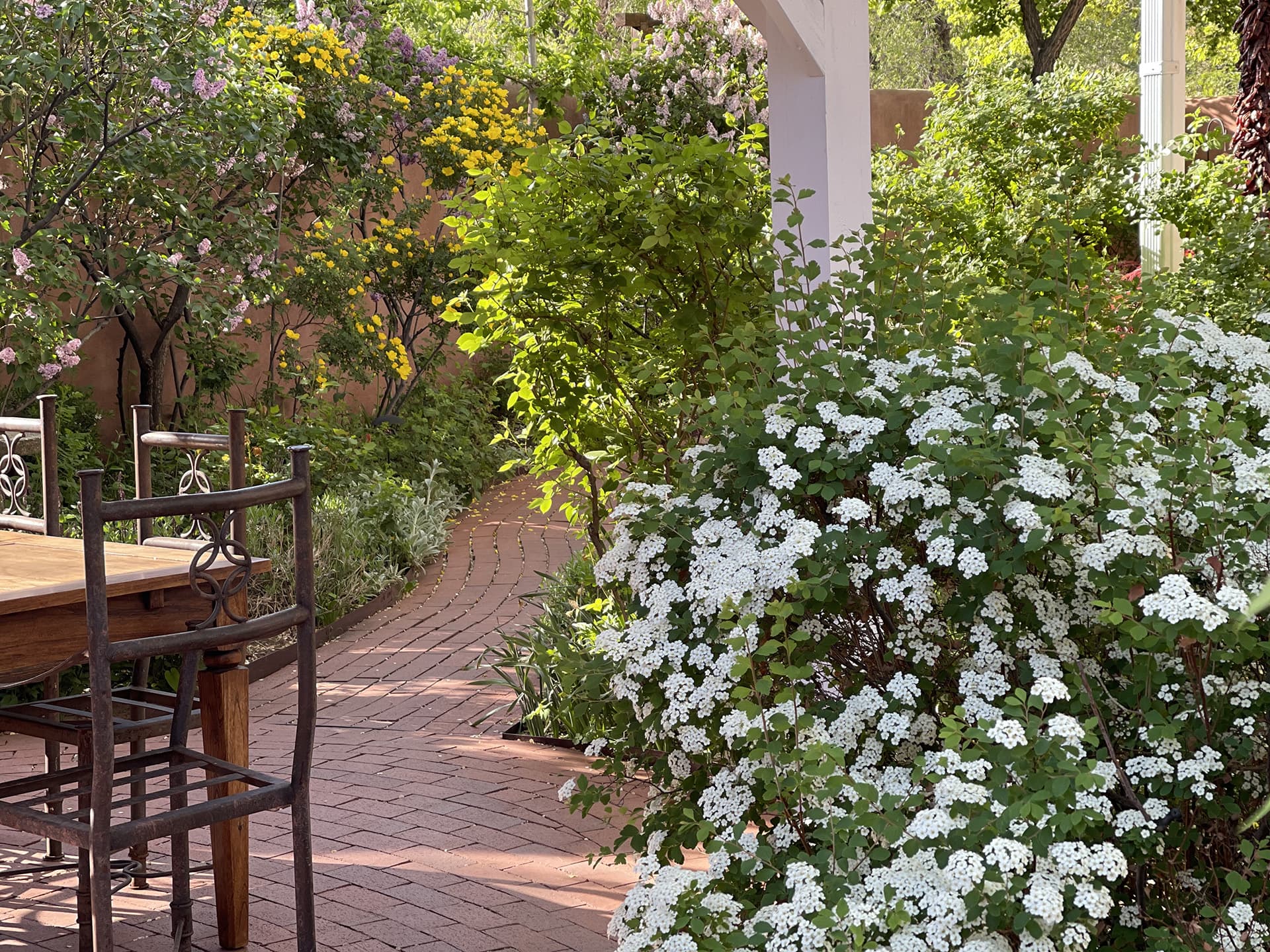 A brick pathway lined with lush greenery and blooming white flowers leads to a wooden table and chairs in a serene garden.