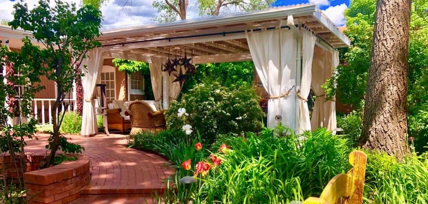 A covered patio at Casa Culinaria with draped white curtains and wicker seating, surrounded by a lush garden of flowers and greenery.