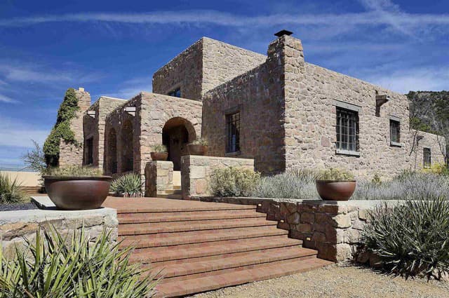 A modern stone house with arched entrances, surrounded by desert landscaping and blue skies.