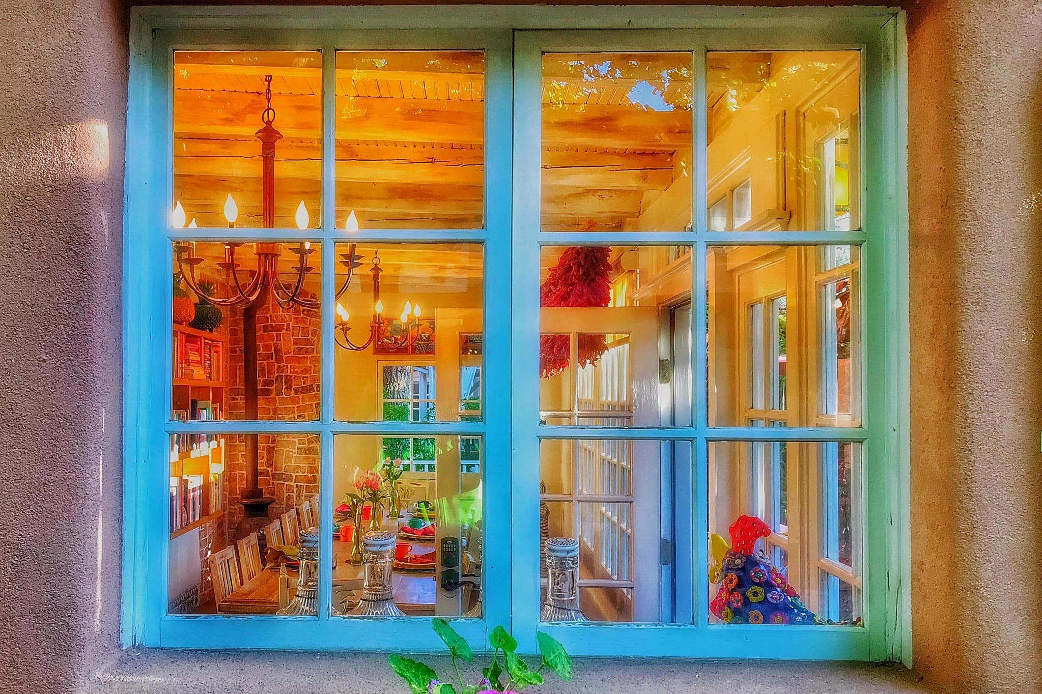 A bright, warmly lit interior view through a blue-framed window, showcasing a dining area with a chandelier and decorative elements.
