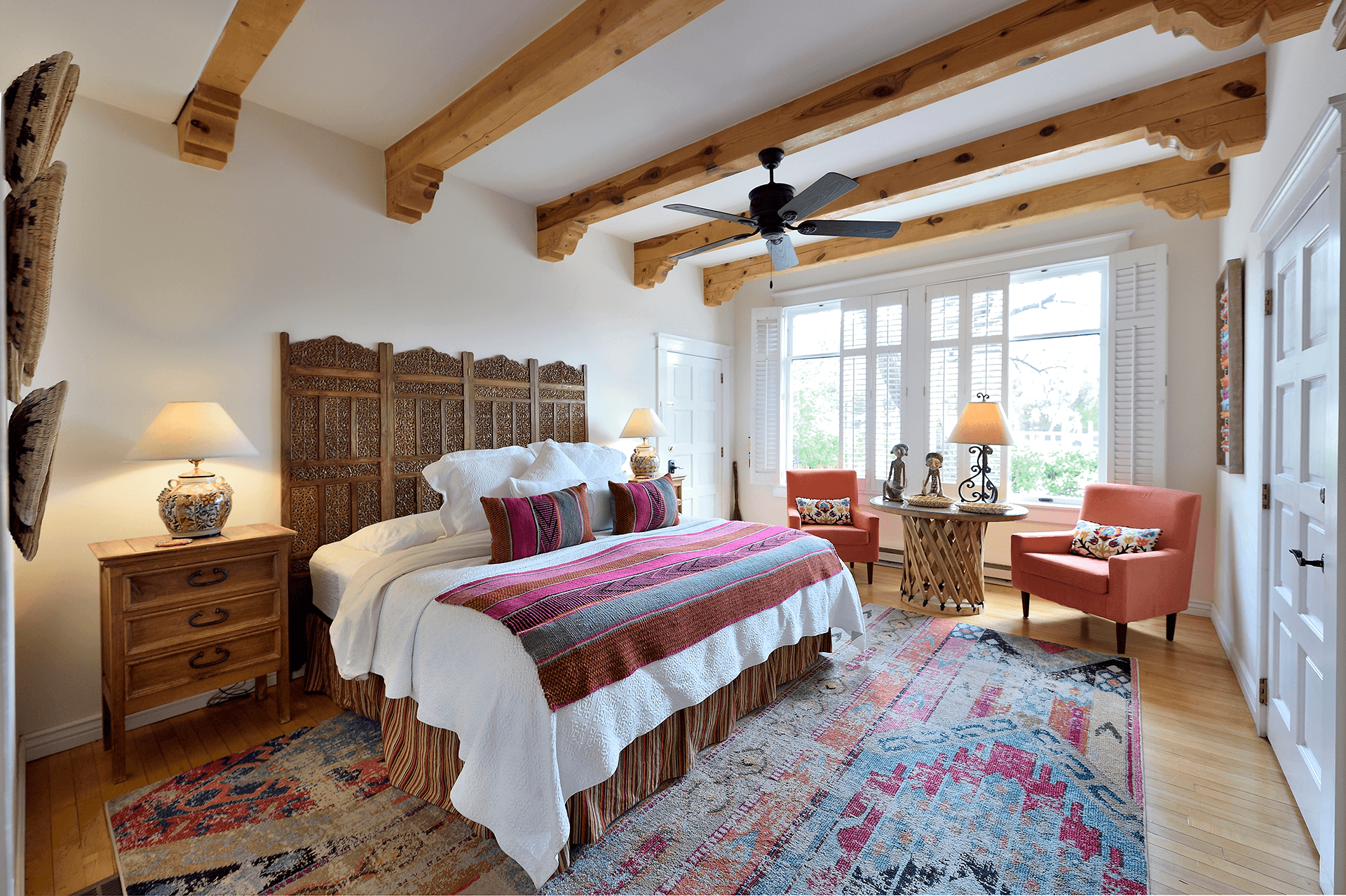 A cozy bedroom featuring a wooden ceiling, a large bed with colorful blankets, and two armchairs next to a window.