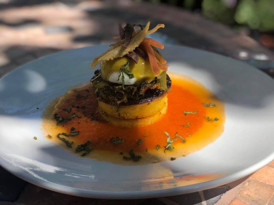 A beautifully plated dish featuring a tower of layered ingredients topped with a poached egg and garnished with colorful strips.