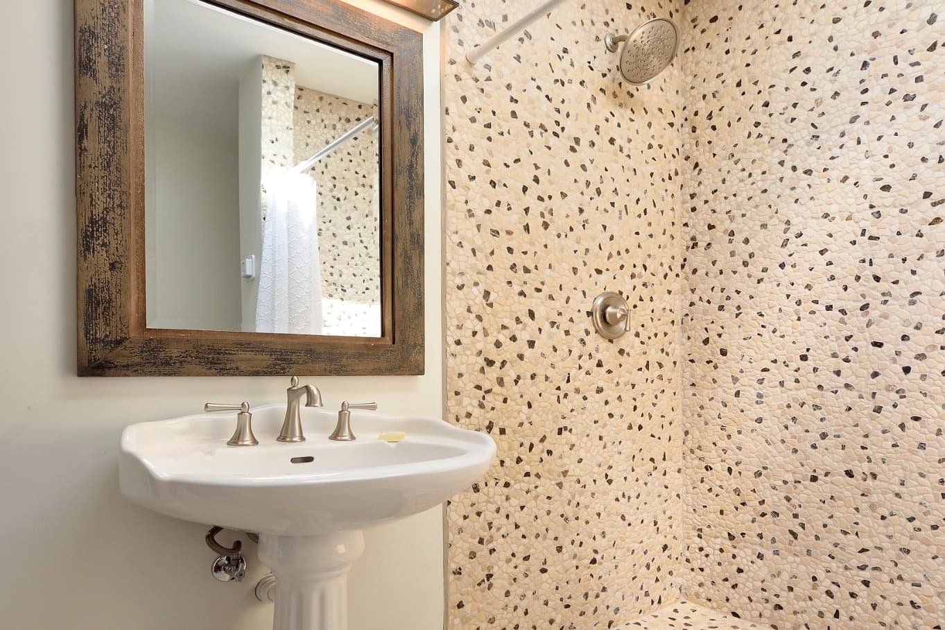 A modern bathroom featuring a pebble stone shower wall, a pedestal sink, and a mirror.