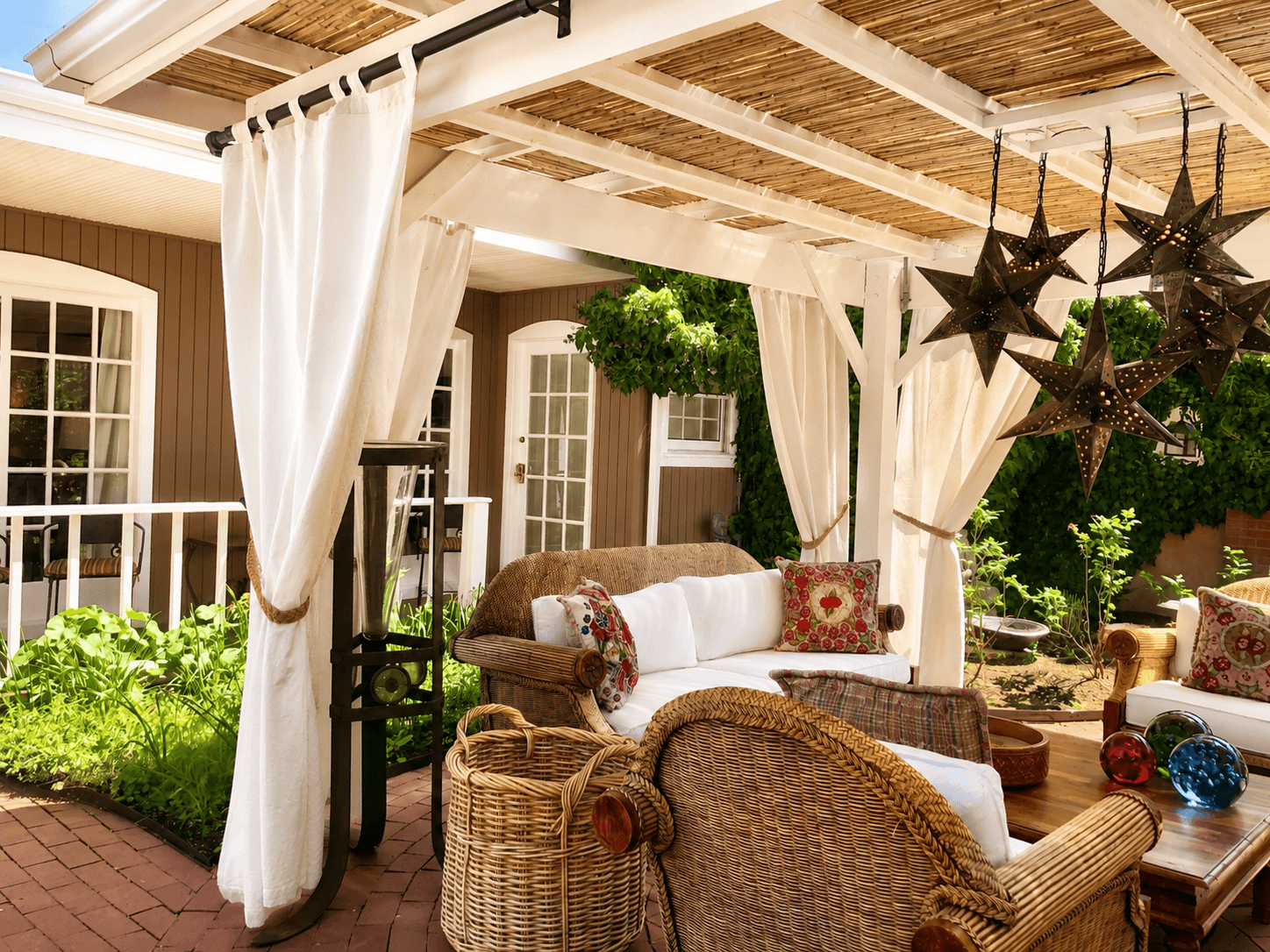 A cozy outdoor seating area with wicker furniture, decorative pillows, and hanging star lanterns under a pergola.