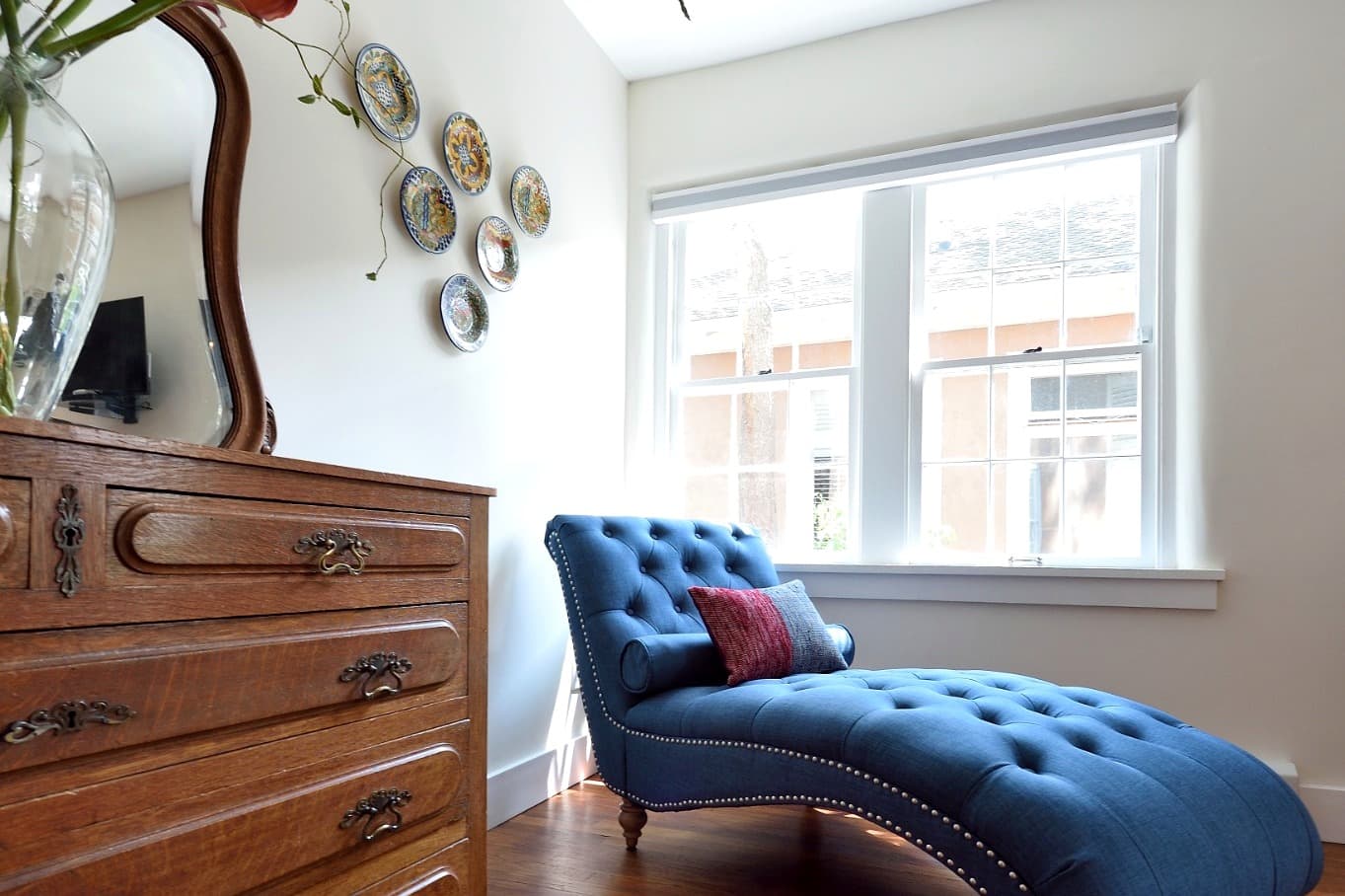 A blue chaise lounge sits beside a wooden dresser, with decorative plates mounted on the wall and sunlight streaming through the window.