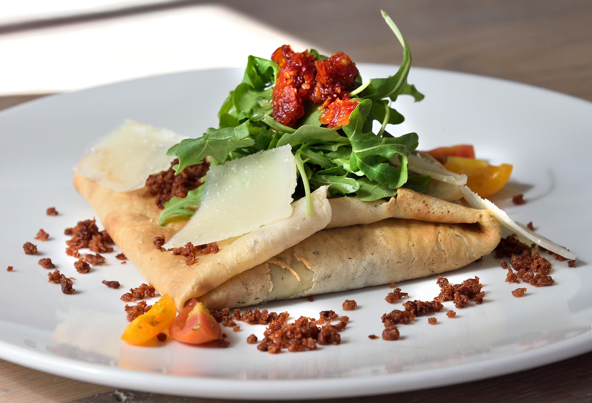 A folded crepe topped with arugula, sun-dried tomatoes, shaved cheese, and crunchy bits on a white plate.