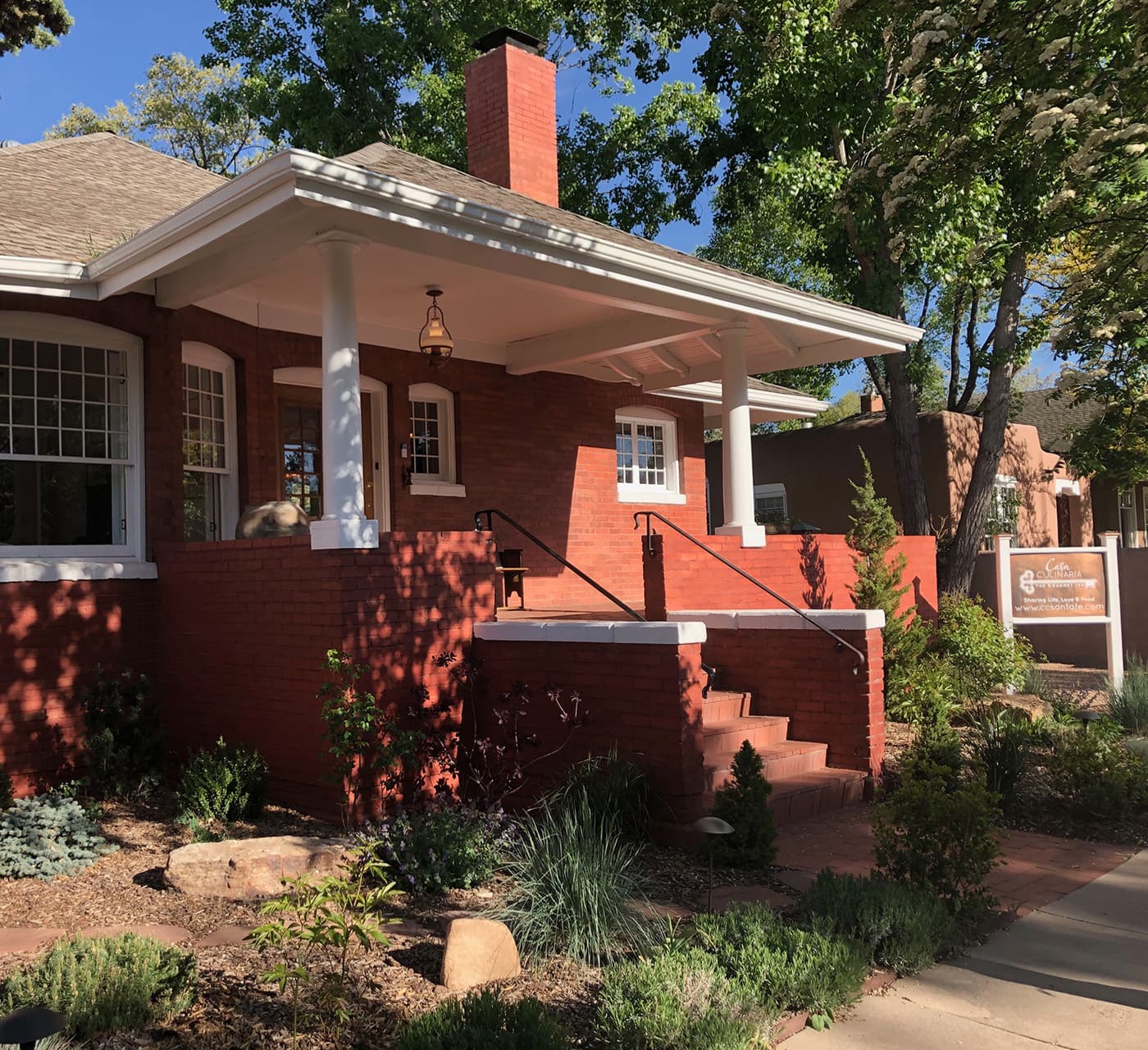 A charming red brick house with a porch, surrounded by greenery and landscaping.