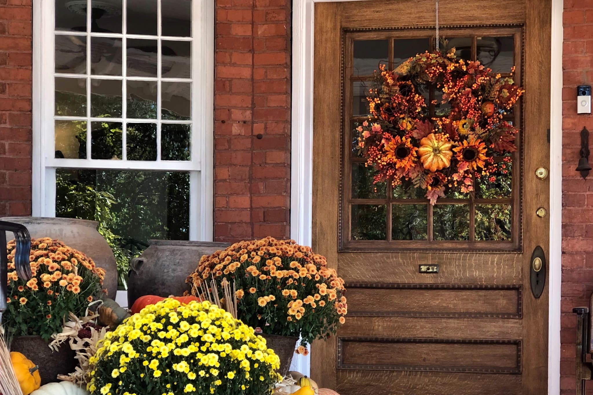 A rustic wooden door adorned with an autumn wreath, surrounded by colorful potted mums and pumpkins.