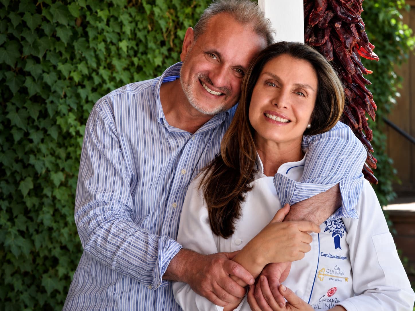 A couple embraces in front of an ivy-covered wall, smiling warmly at the camera.