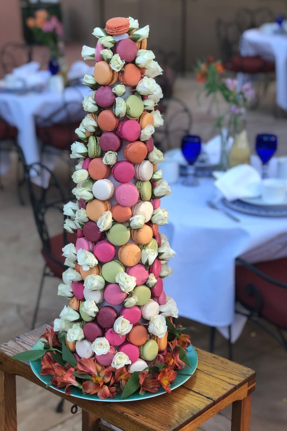 A colorful tower of macarons adorned with white roses, displayed on a wooden table.