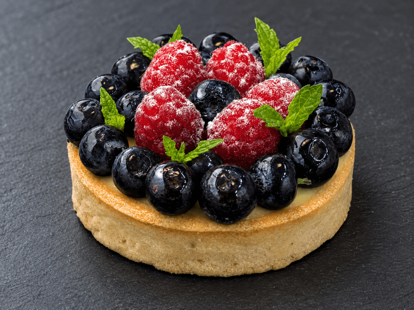 A round fruit tart topped with raspberries, blueberries, and fresh mint on a dark background.