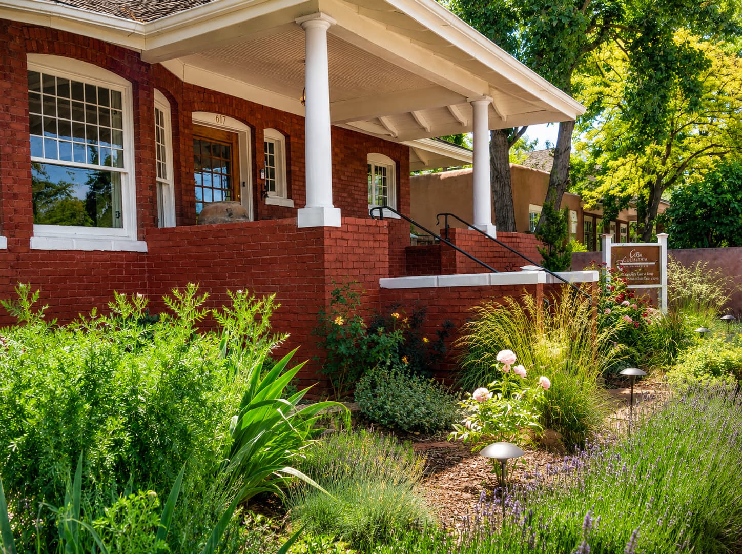 A charming brick house with a welcoming porch, surrounded by lush greenery and colorful flowers.
