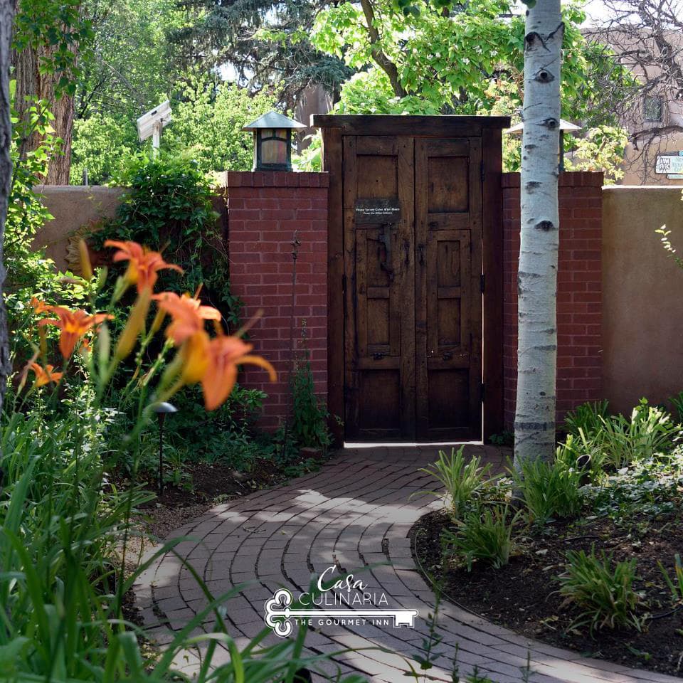 A wooden door set in a brick wall is surrounded by lush greenery and blooming flowers along a curved path.