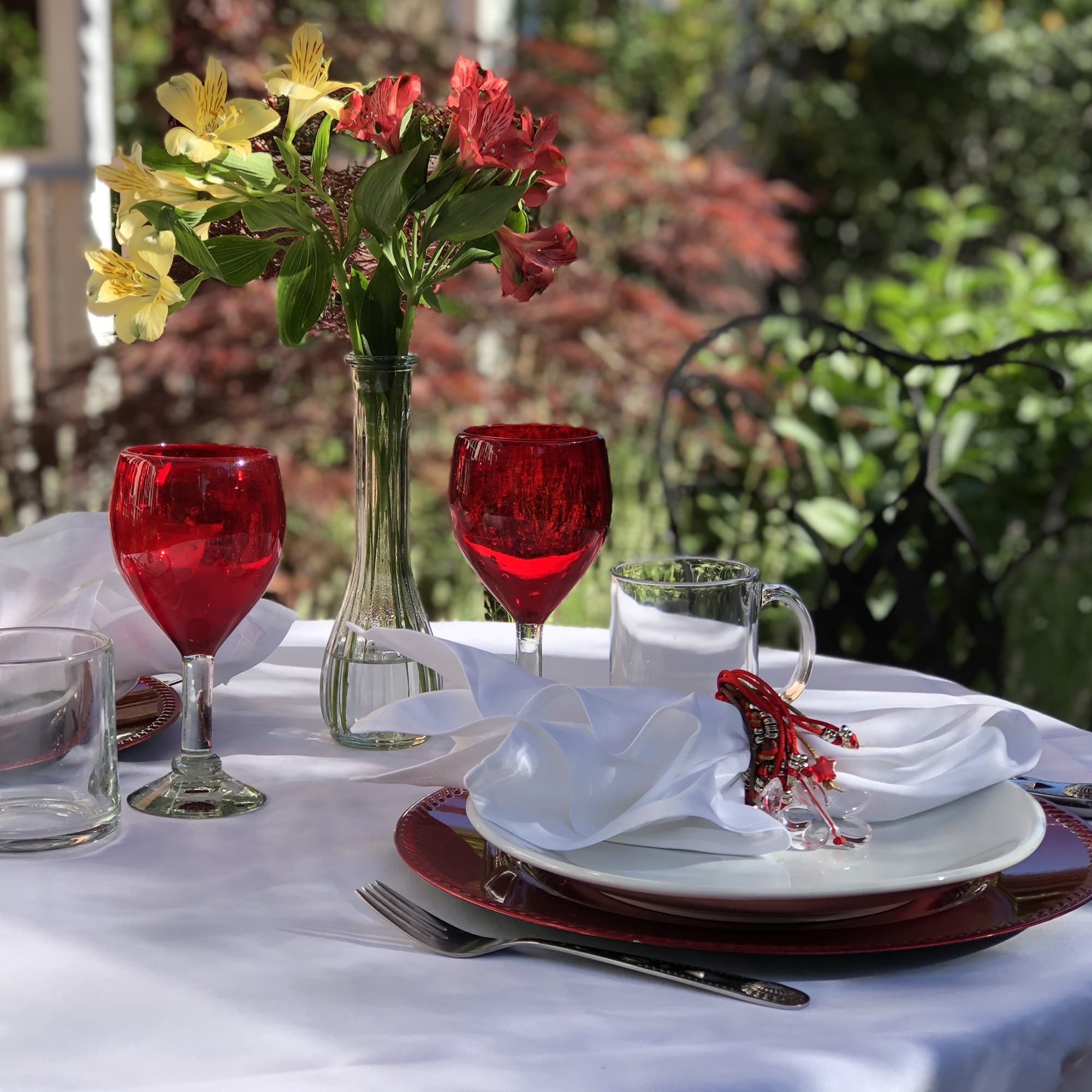 A beautifully set outdoor dining table with red glasses, white napkins, and a floral centerpiece.