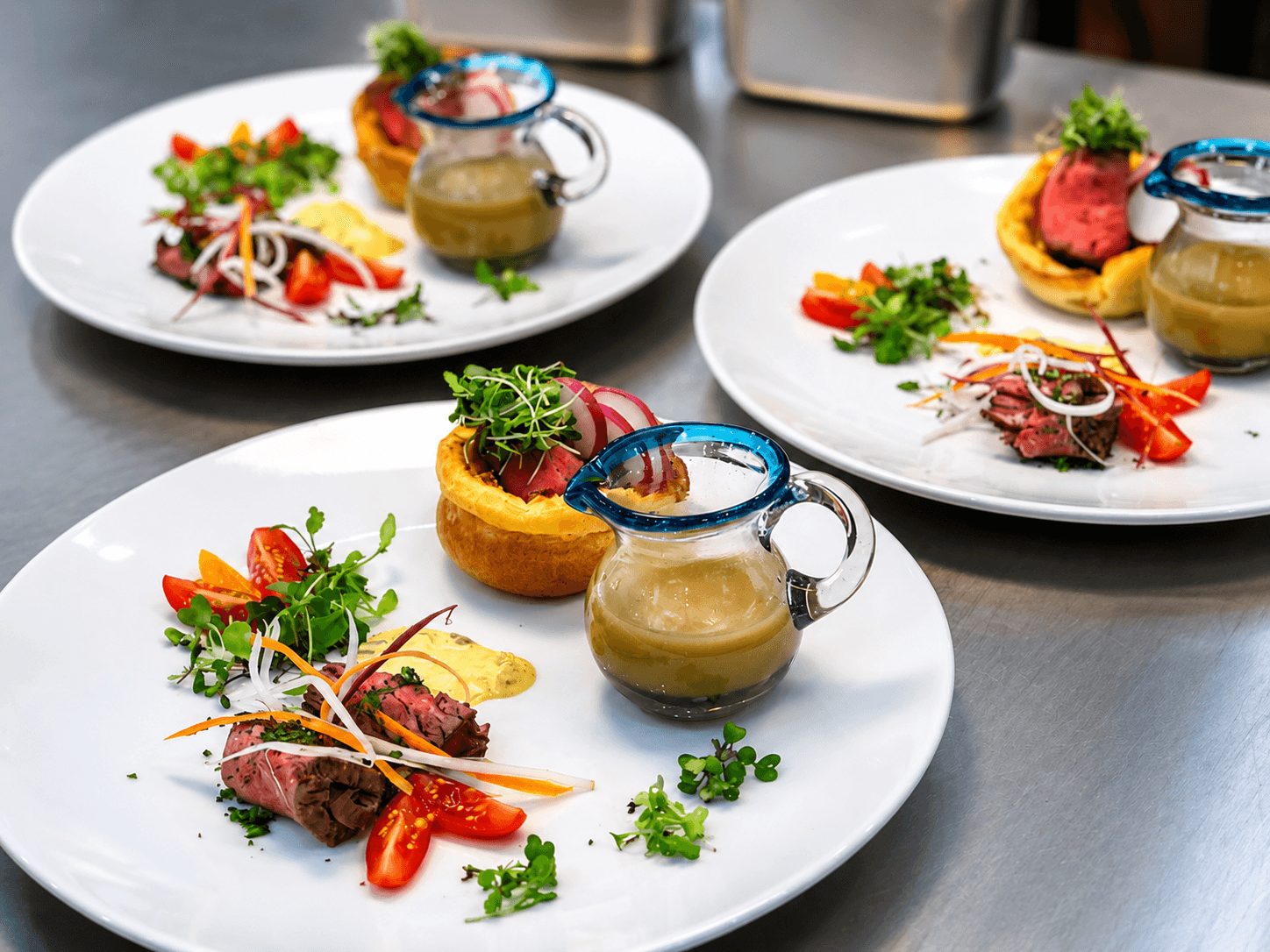 Three elegantly plated dishes featuring sliced beef, colorful vegetables, and a small jug of sauce.
