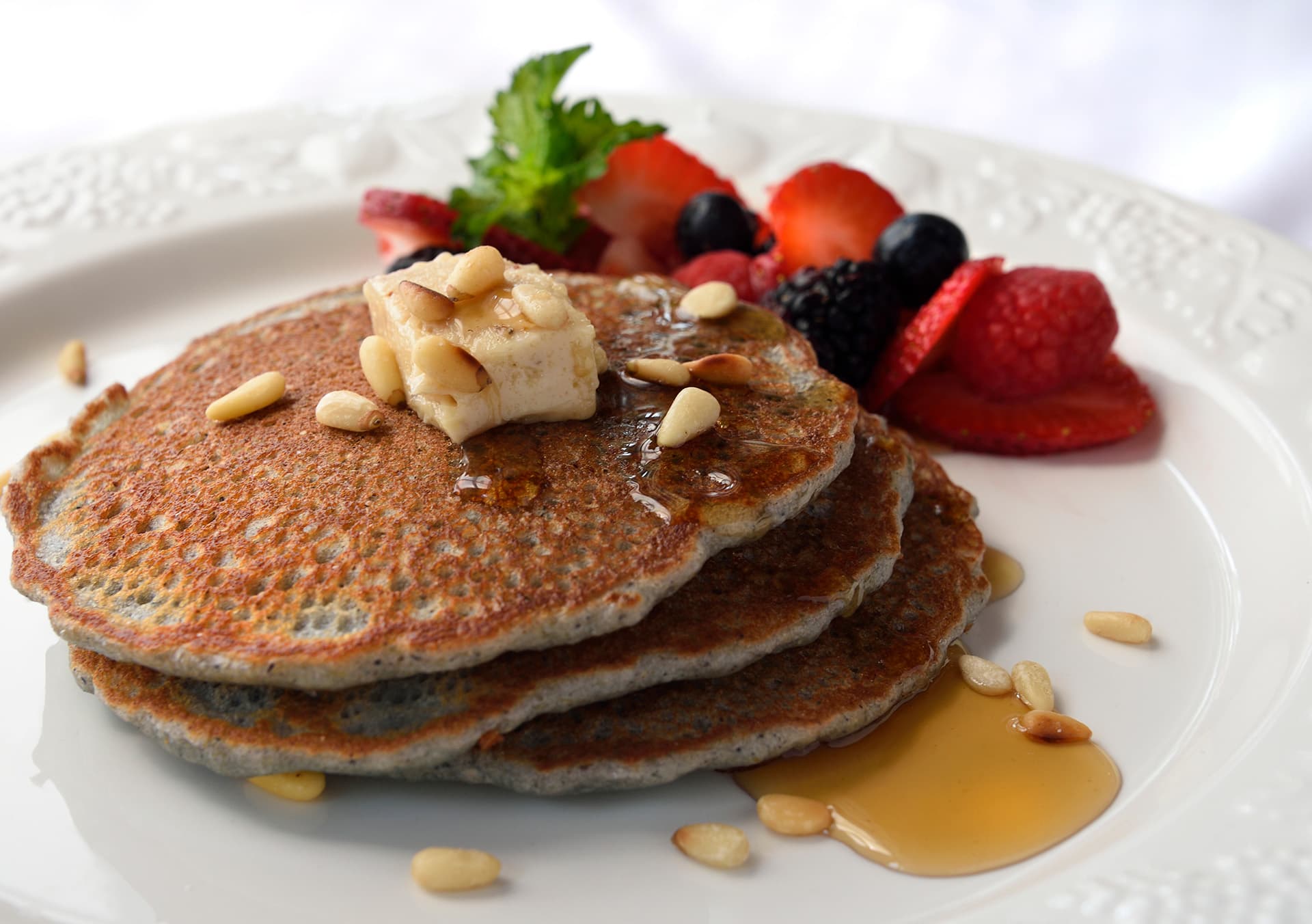 Three pancakes topped with a pat of butter, pine nuts, and syrup, accompanied by fresh berries on the side.
