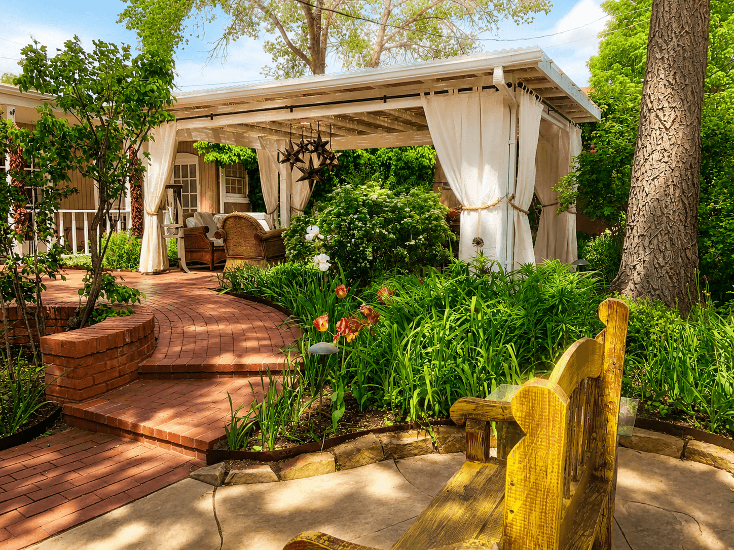 A charming outdoor patio with white curtains, surrounded by lush greenery and colorful flowers, features comfortable seating and a winding brick pathway.