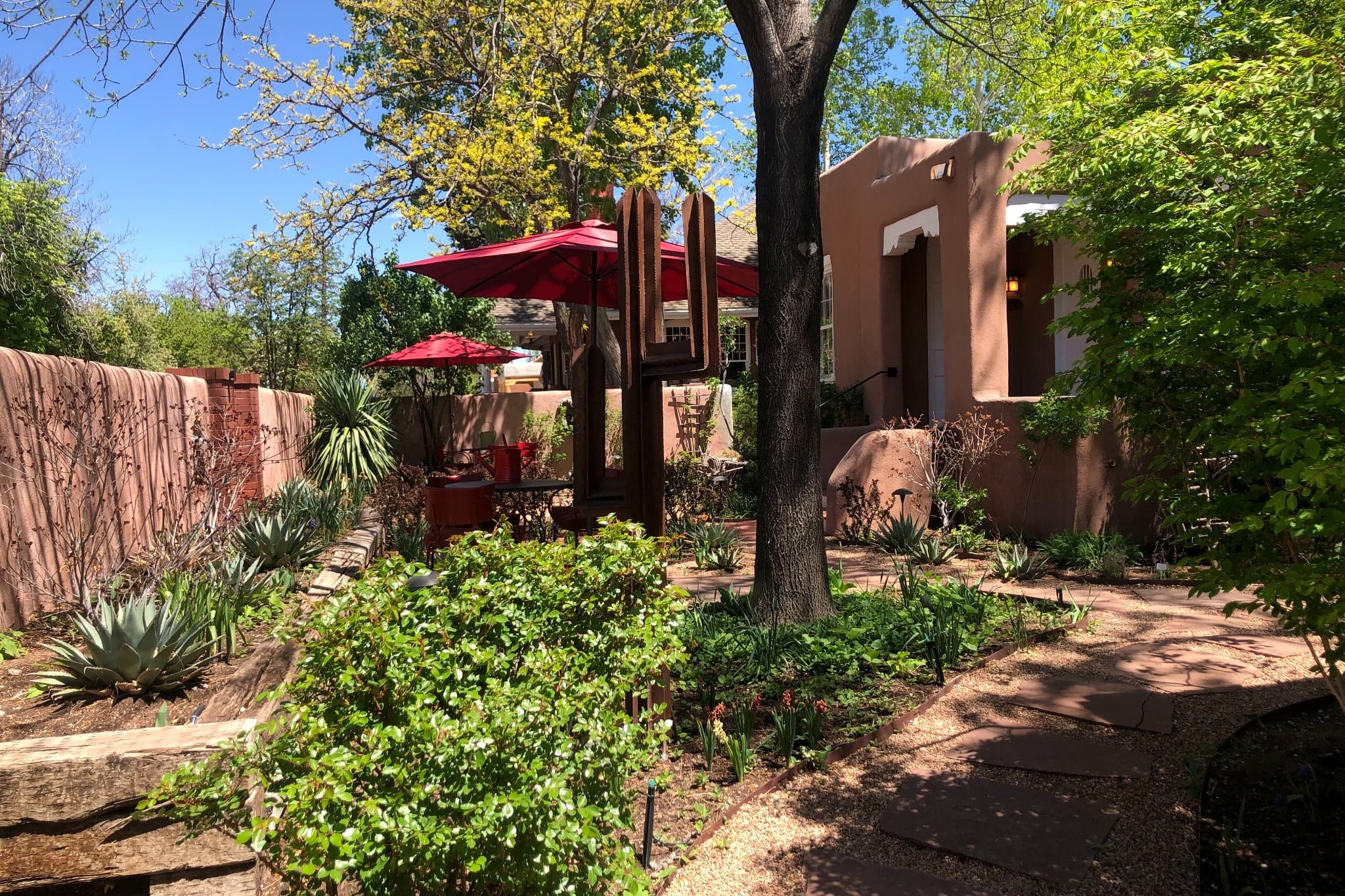 A vibrant garden with lush greenery and a shaded seating area beside a brown adobe building.