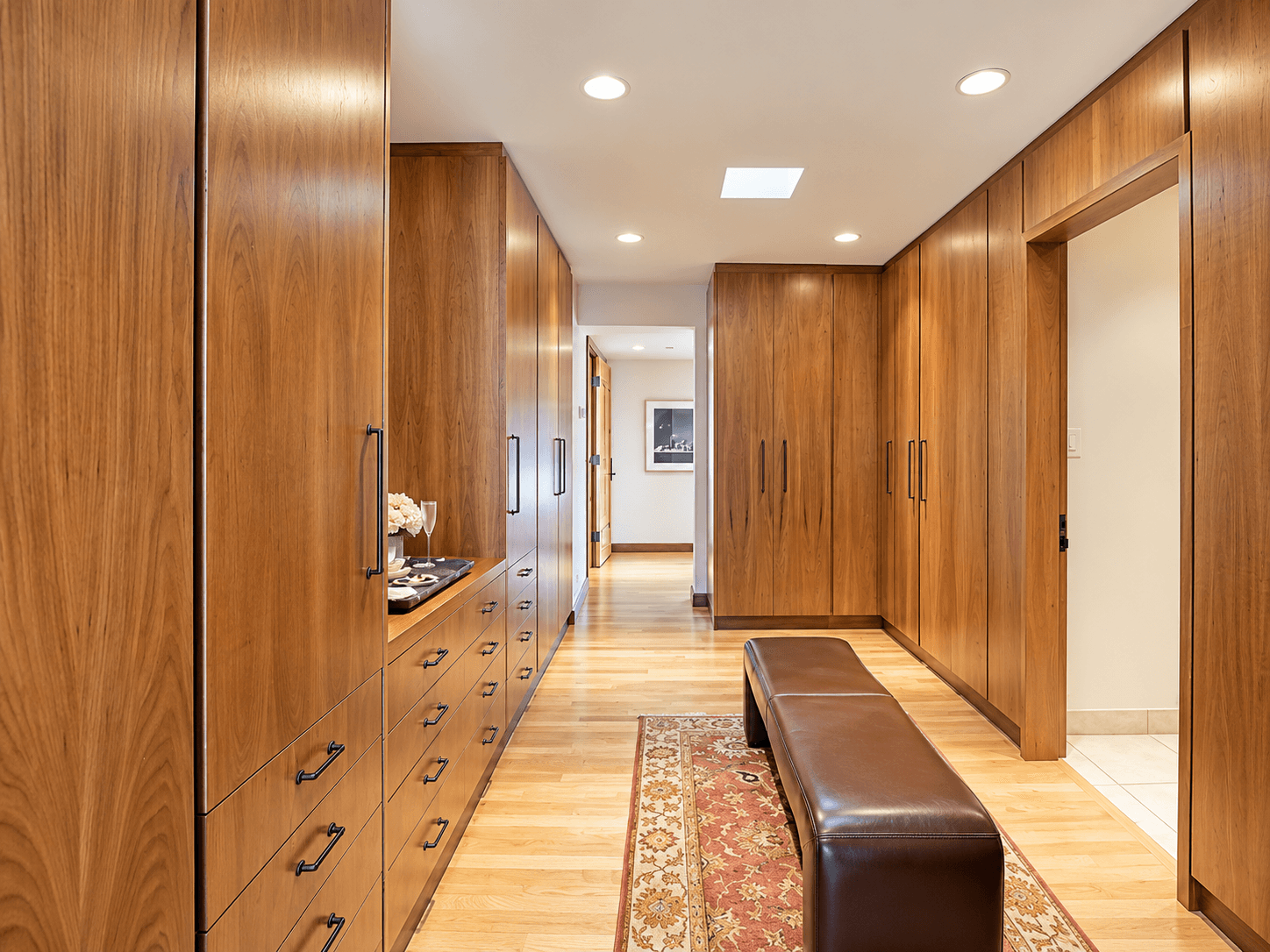 A spacious hallway featuring wooden cabinetry, a leather bench, and warm lighting.