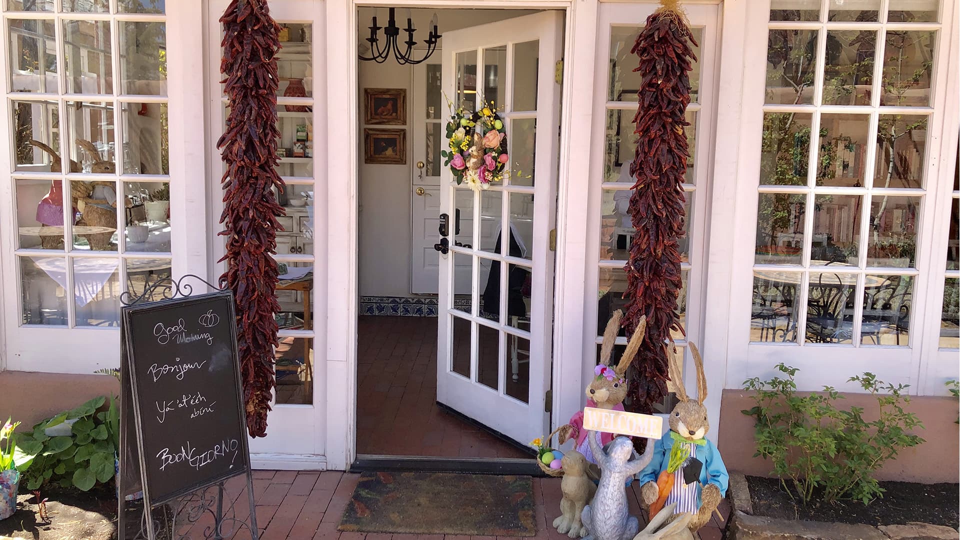 A charming restaurant entrance adorned with red peppers, a floral wreath, and decorative rabbits, with a chalkboard menu nearby.
