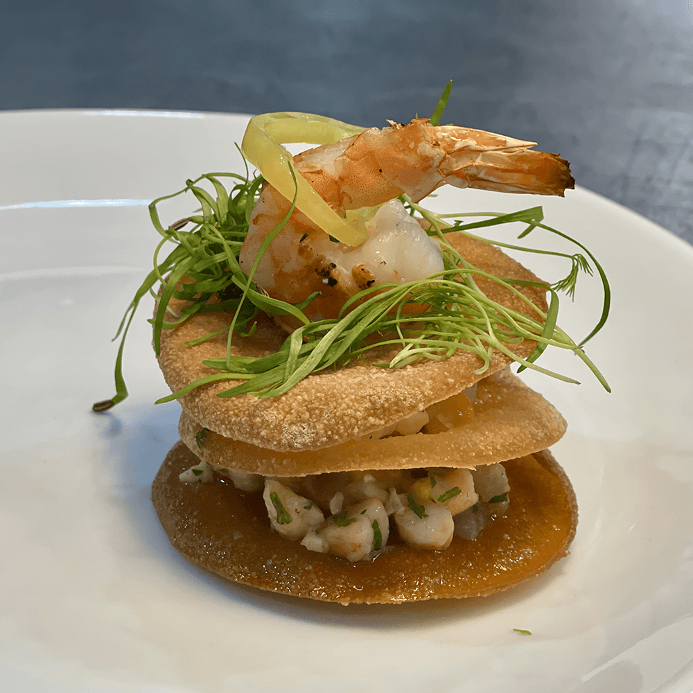 A stacked appetizer featuring crispy flatbreads topped with shrimp, microgreens, and garnish.