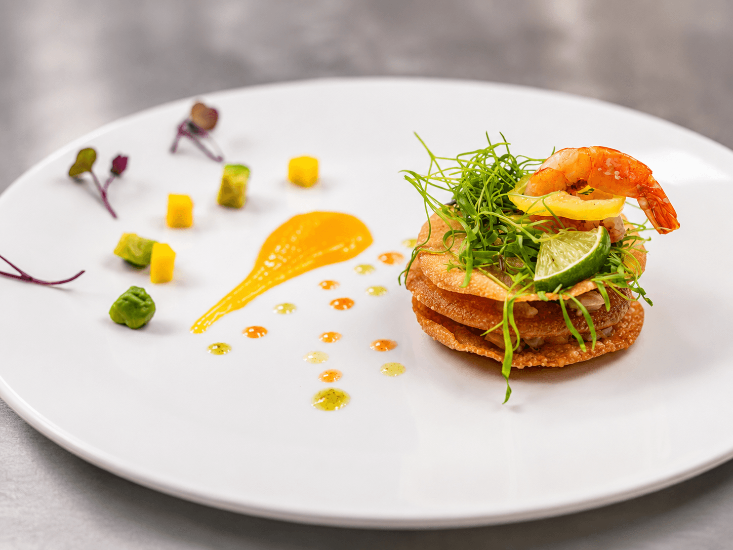 A beautifully plated dish featuring a stack of crispy layers topped with shrimp, fresh herbs, and garnishes, accompanied by colorful sauces and vegetables.