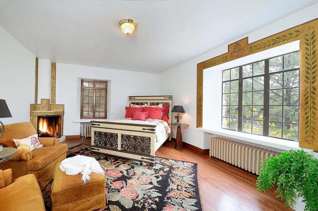 Cozy bedroom featuring a decorative bed, a fireplace, and large windows with a colorful rug.