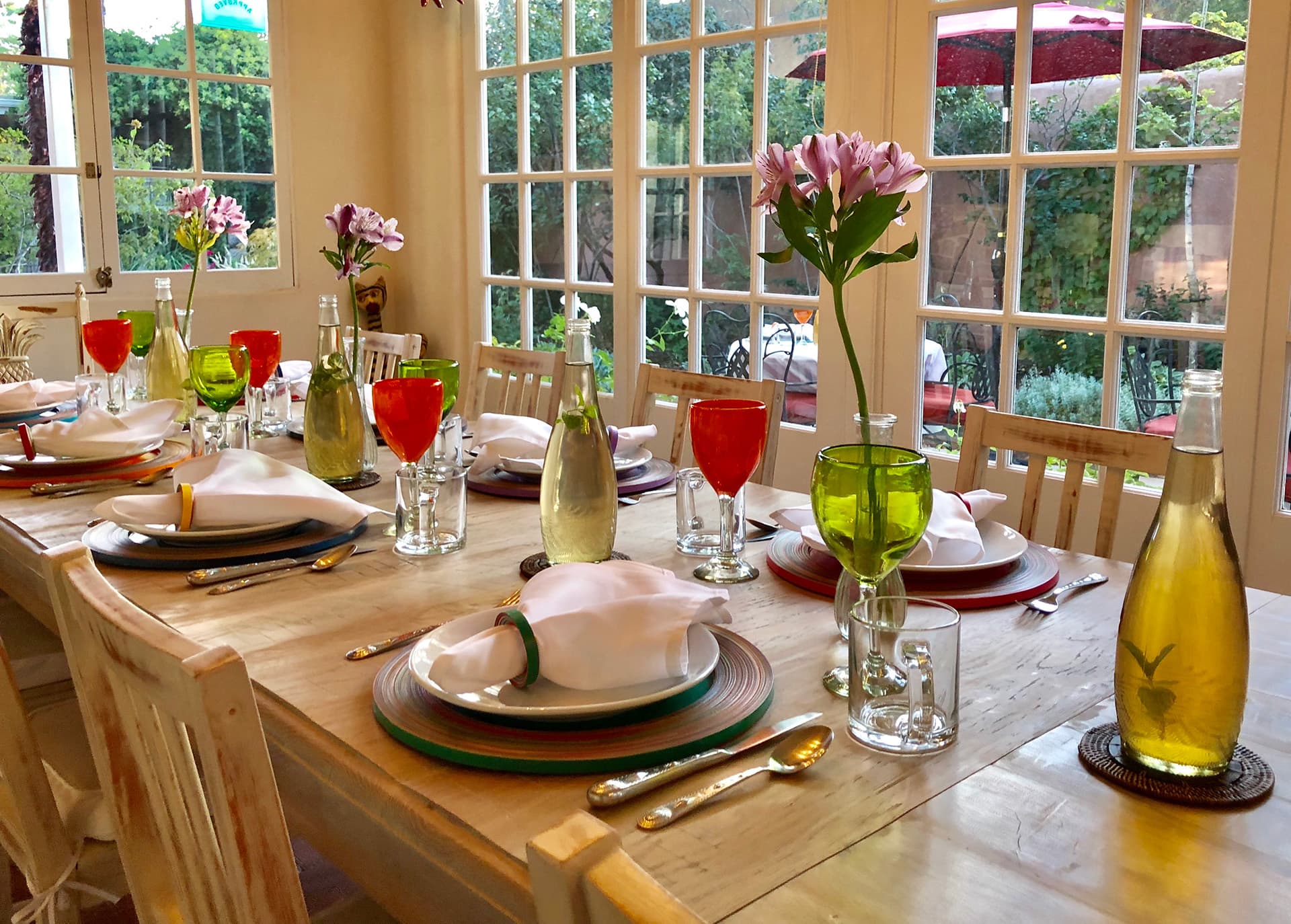 A beautifully set dining table with colorful glasses, folded napkins, and floral centerpieces, beside large windows.