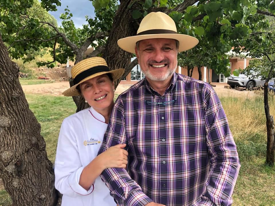 A man and a woman, both smiling, pose together under a tree, wearing hats.