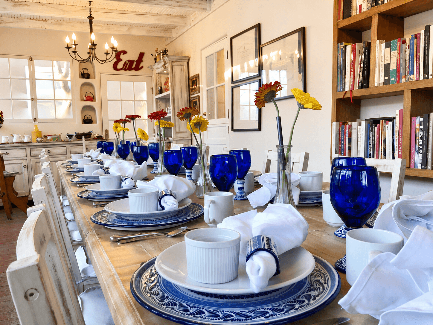 A beautifully set dining table with blue dishes, white napkins, and colorful flowers, surrounded by bookshelves and decorative items.