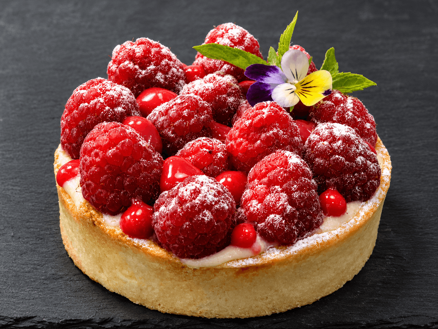 A delicious raspberry tart topped with fresh raspberries, edible flowers, and a dusting of powdered sugar.