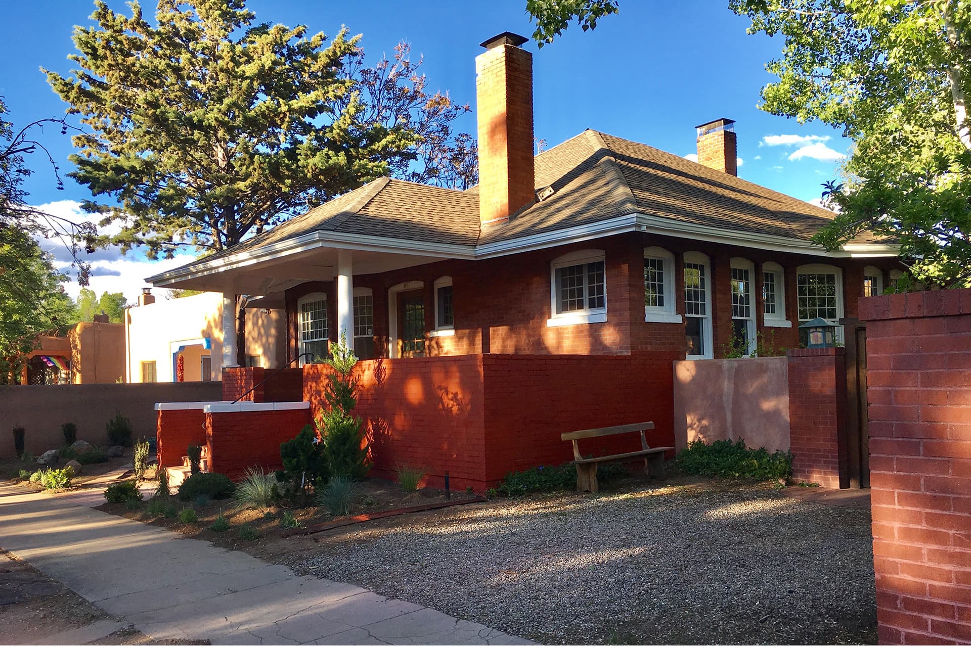 A red brick house with a front porch and large windows, surrounded by greenery and a pathway.