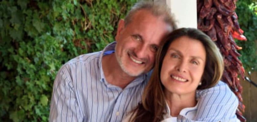 A smiling couple embraces in front of greenery and hanging red peppers.