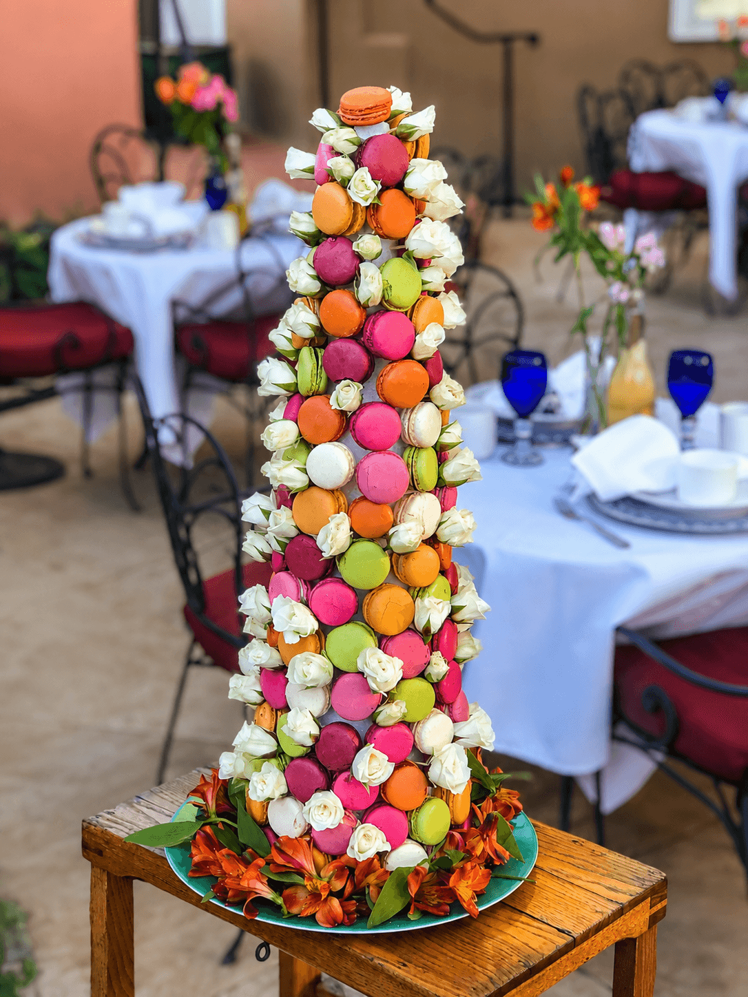 A tall, colorful tower of macarons surrounded by white roses, displayed on a rustic wooden table in a quaint outdoor dining setting.